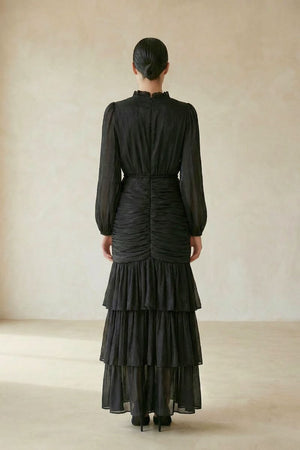 Back view of a model in a black long-sleeve maxi dress featuring ruched detailing and layered tiered skirt, shown against a soft beige background.