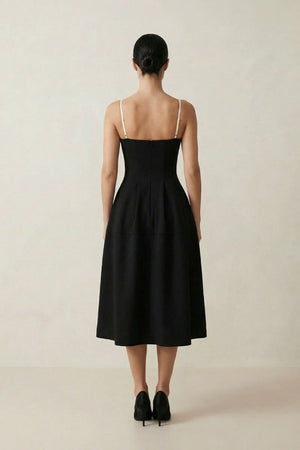 Back view of the black midi dress with adjustable ivory straps, a fitted waist, and a graceful A-line skirt shown against a neutral studio backdrop.