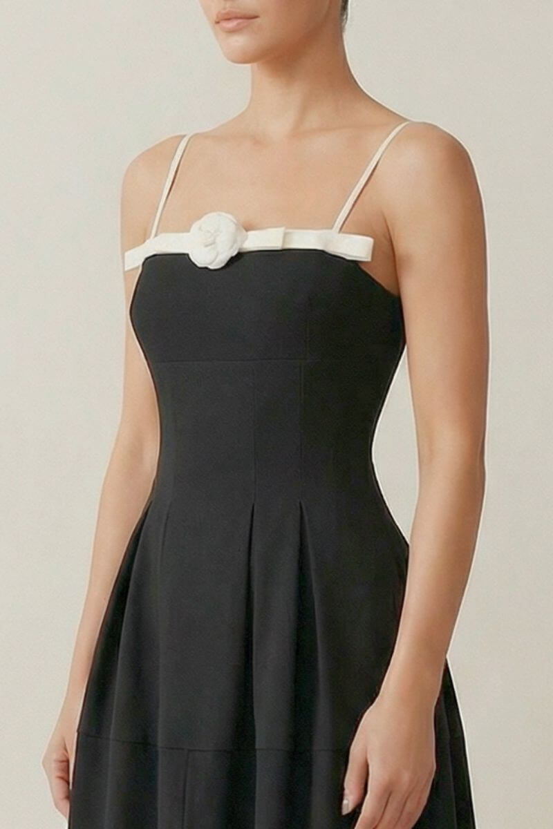 Close-up of the black midi dress showing the fitted bodice, ivory satin trim, and small white rosette accent across the neckline.