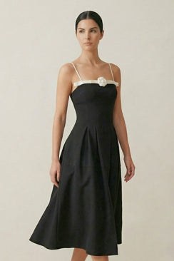 Model wearing a black midi dress with thin ivory straps and a white rosette at the neckline, styled in a clean studio setting for a refined, minimalist look.