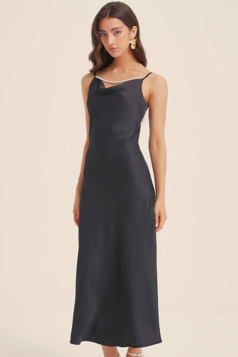 Woman wearing a black satin maxi slip dress featuring a draped neckline and pearl-trim strap detail, styled with gold earrings and white heels for an elegant evening look.