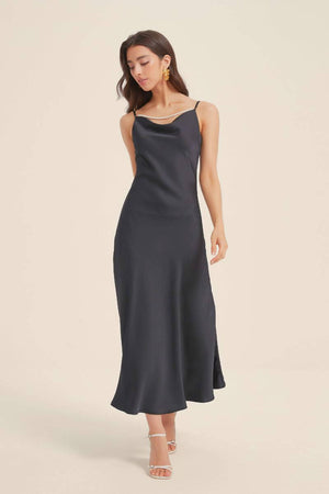 Model wearing a full-length black satin slip dress with a soft cowl neckline and pearl strap accent, creating a sleek, timeless silhouette for evening occasions.