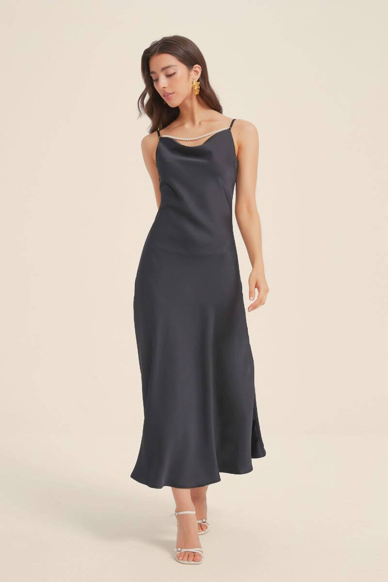 Model wearing a full-length black satin slip dress with a soft cowl neckline and pearl strap accent, creating a sleek, timeless silhouette for evening occasions.