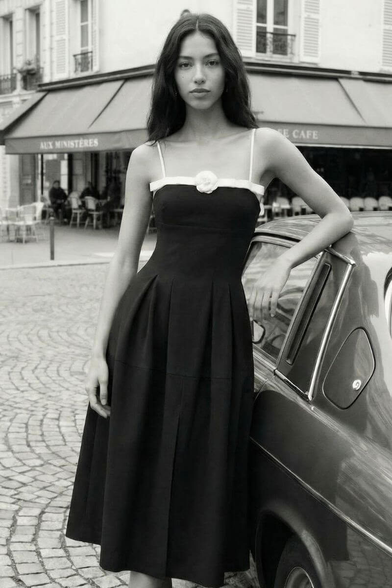 A woman leans against a vintage car on a Paris street wearing a black midi dress with thin straps, a contrast trim neckline, and a single rosette detail at the bust.