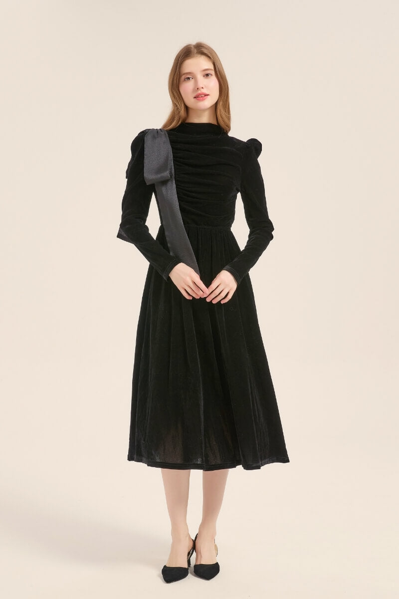 A woman in a black velvet long-sleeve dress showing soft ruching and a sculpted fit with an elegant satin shoulder bow.