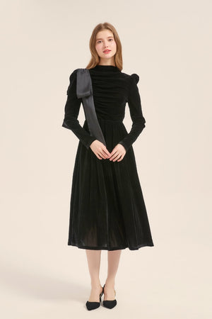 A woman in a black velvet long-sleeve dress showing soft ruching and a sculpted fit with an elegant satin shoulder bow.