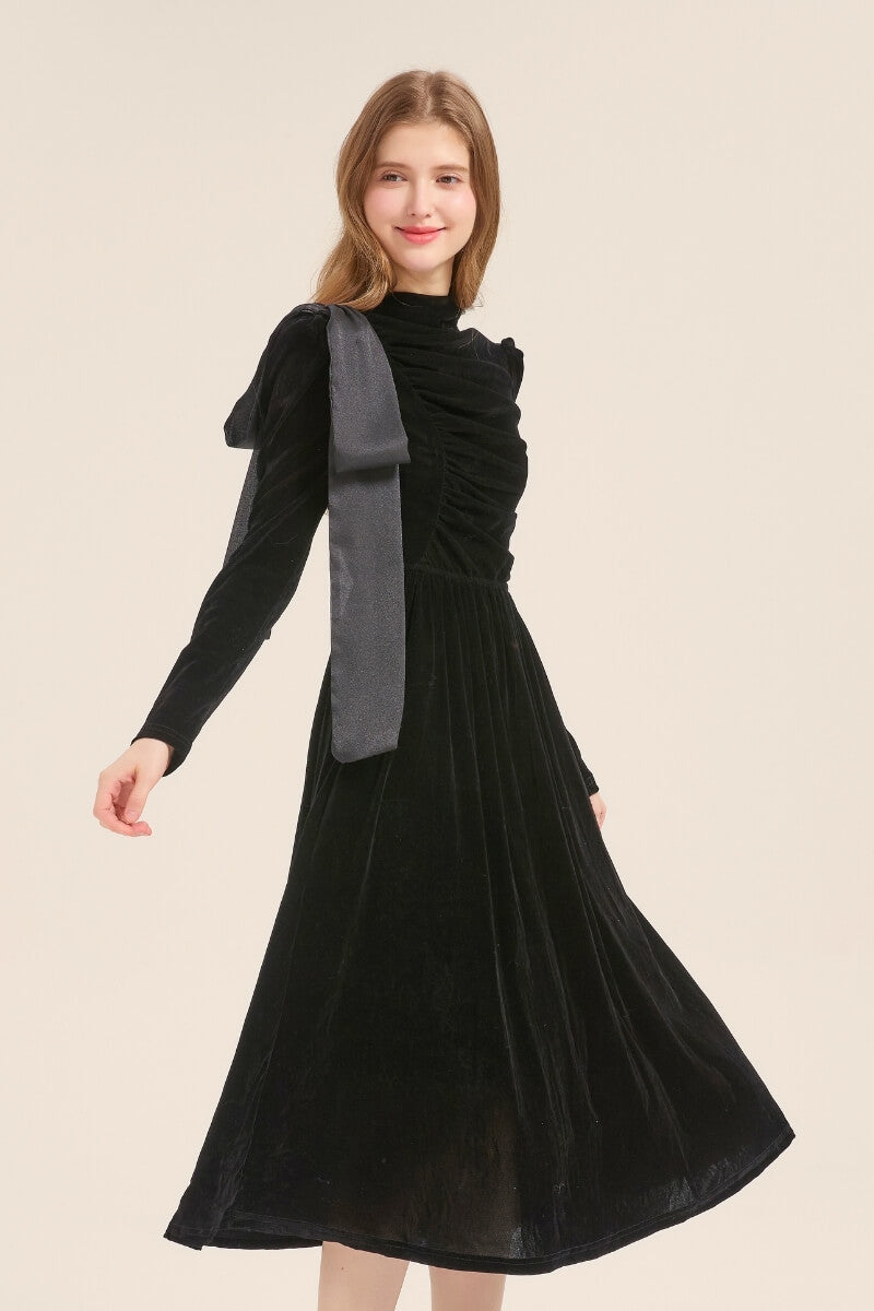 Model smiling in a long-sleeve black velvet dress with ruched texture and asymmetric bow shoulder tie, styled for an evening look.