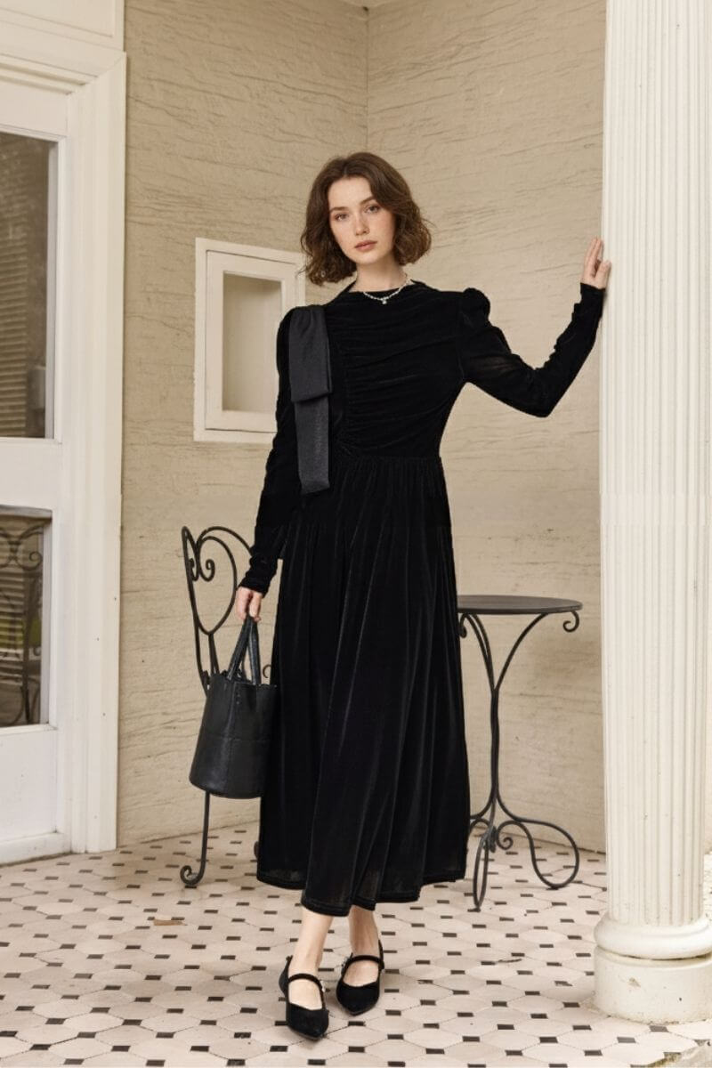Model standing on a tiled veranda wearing a black velvet maxi dress with long sleeves, ruched bodice, and shoulder bow detail, paired with black Mary Jane heels and a bucket bag beside an iron café chair.