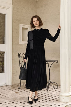 Model standing on a tiled veranda wearing a black velvet maxi dress with long sleeves, ruched bodice, and shoulder bow detail, paired with black Mary Jane heels and a bucket bag beside an iron café chair.