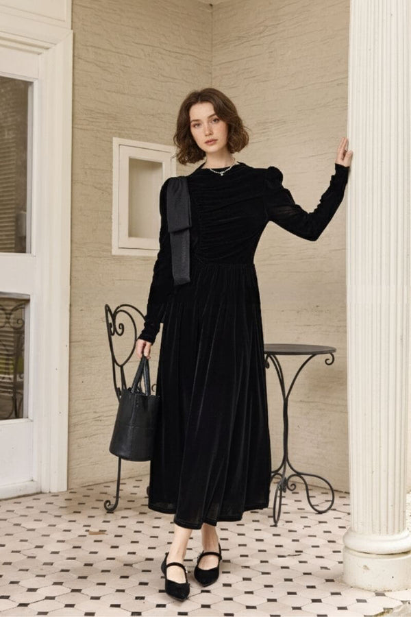 Model standing on a tiled veranda wearing a black velvet maxi dress with long sleeves, ruched bodice, and shoulder bow detail, paired with black Mary Jane heels and a bucket bag beside an iron café chair.