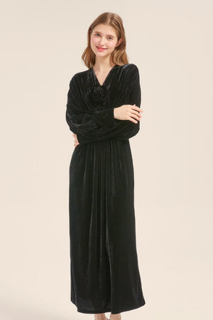 Model wearing a long black velvet maxi dress with deep V-neckline, gathered waist, and velvet rose appliqué detail.