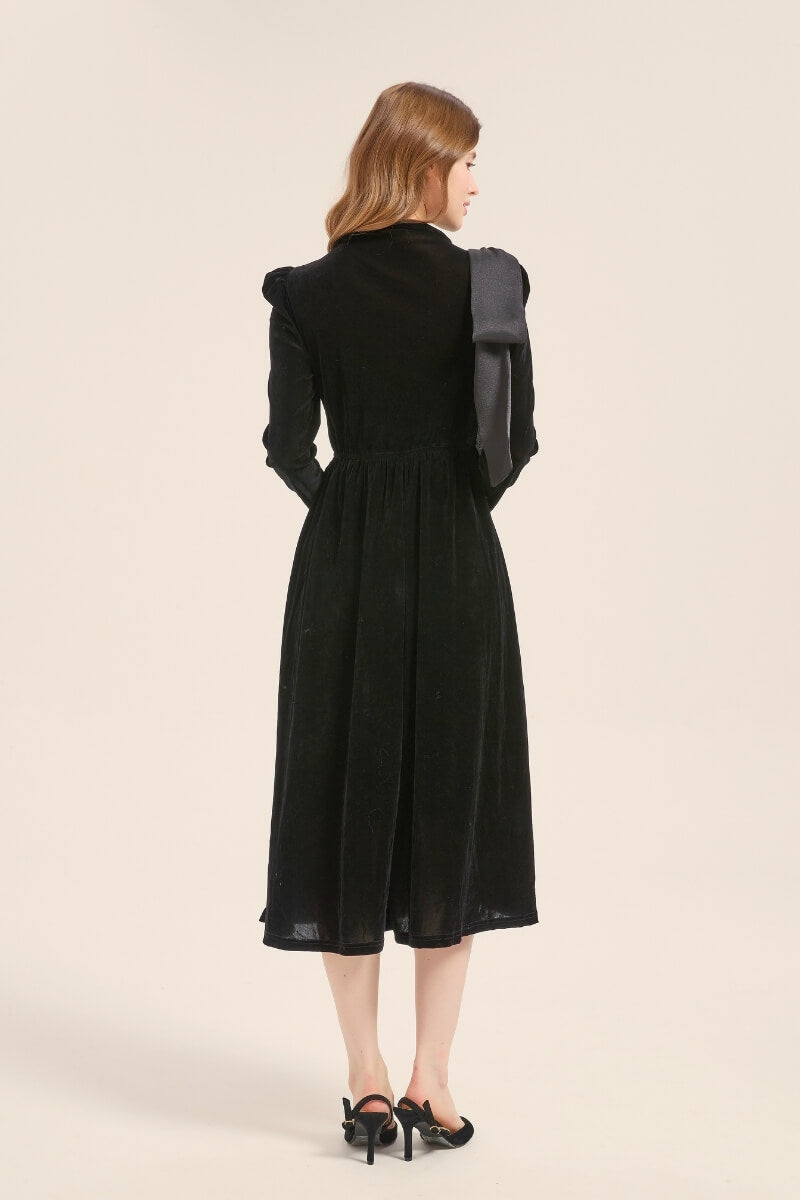 Back view of a black velvet midi dress with long sleeves and soft flare hem, featuring a large bow accent draped from the shoulder.