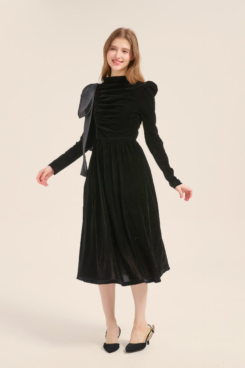 Model posing in a full-length view of a black velvet midi dress with puff shoulders, ruched front, and statement satin bow detail.