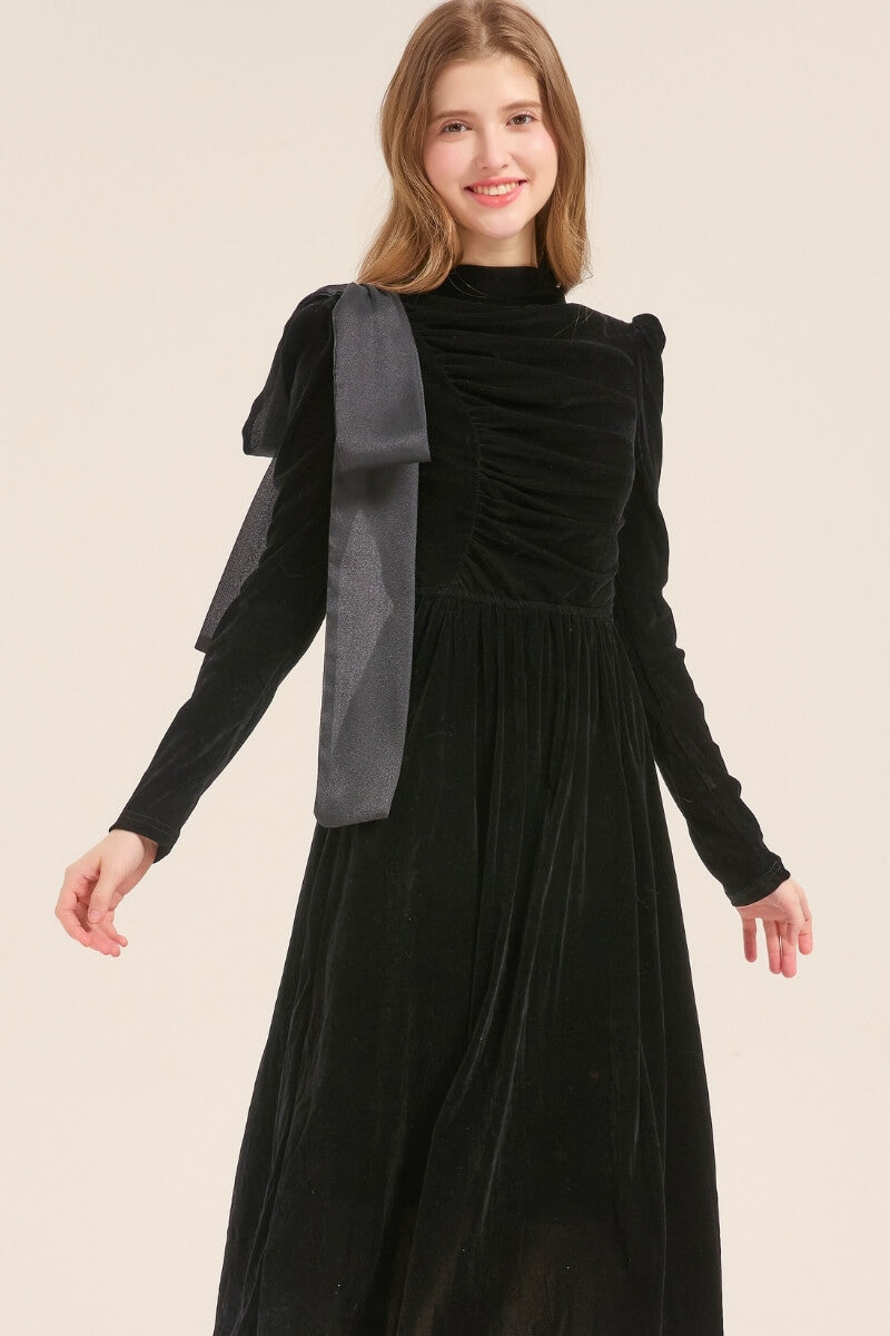 Model wearing a black velvet midi dress featuring long sleeves, ruched bodice, and an oversized bow detail on the shoulder, styled with black slingback heels.