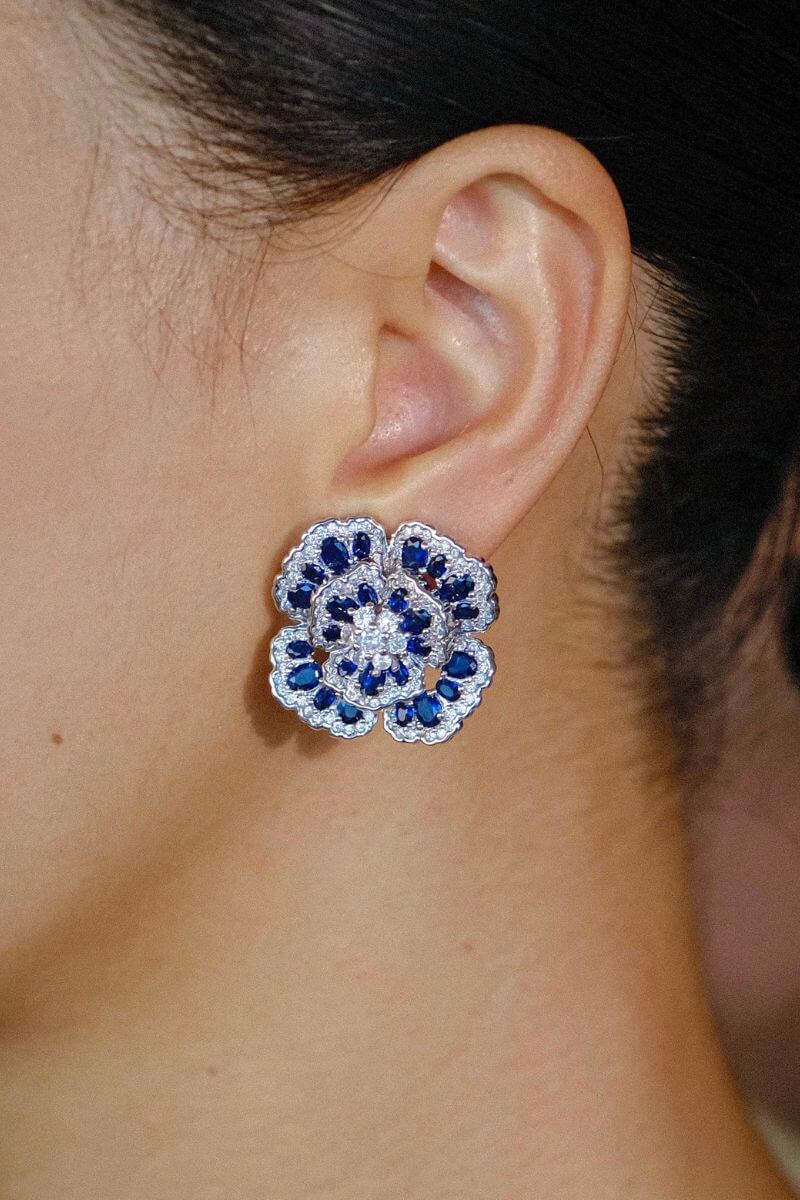 Close-up view of a blue floral gemstone stud earring worn on the ear, featuring layered petal design with deep blue stones and sparkling clear accents.