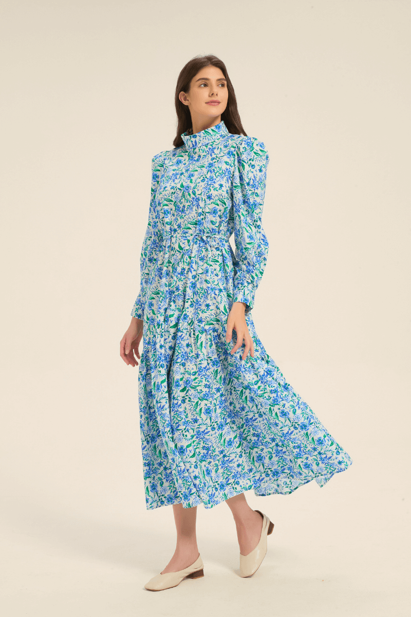 Model walking in a blue floral high neck midi dress with puffed shoulders and tiered skirt, side-front view.