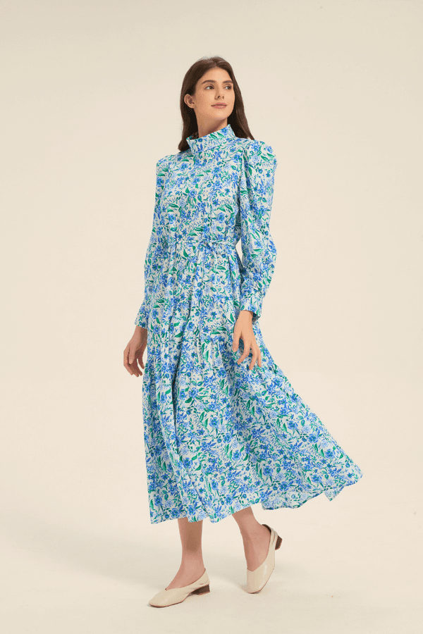 Model walking in a blue floral high neck midi dress with puffed shoulders and tiered skirt, side-front view.