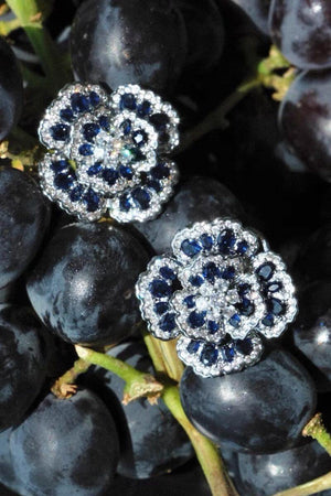 Blue floral gemstone stud earrings styled on dark grapes, creating a rich editorial still life that highlights the deep blue stones and crystal detailing.