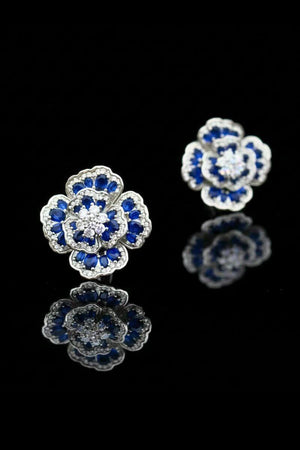 Blue flower gemstone stud earrings photographed on a glossy black surface, creating a mirrored reflection that emphasizes the intricate petal setting.