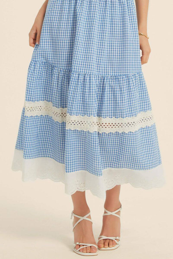 Close-up of a blue gingham midi skirt with scalloped white lace panels and embroidered hem, styled with white strappy heels.