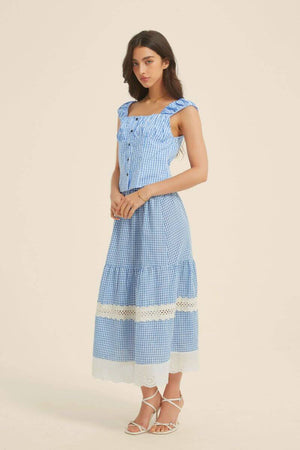 Side view of a woman in a blue gingham crop top and tiered maxi skirt with lace panel details, styled with white heels and minimal gold jewelry.