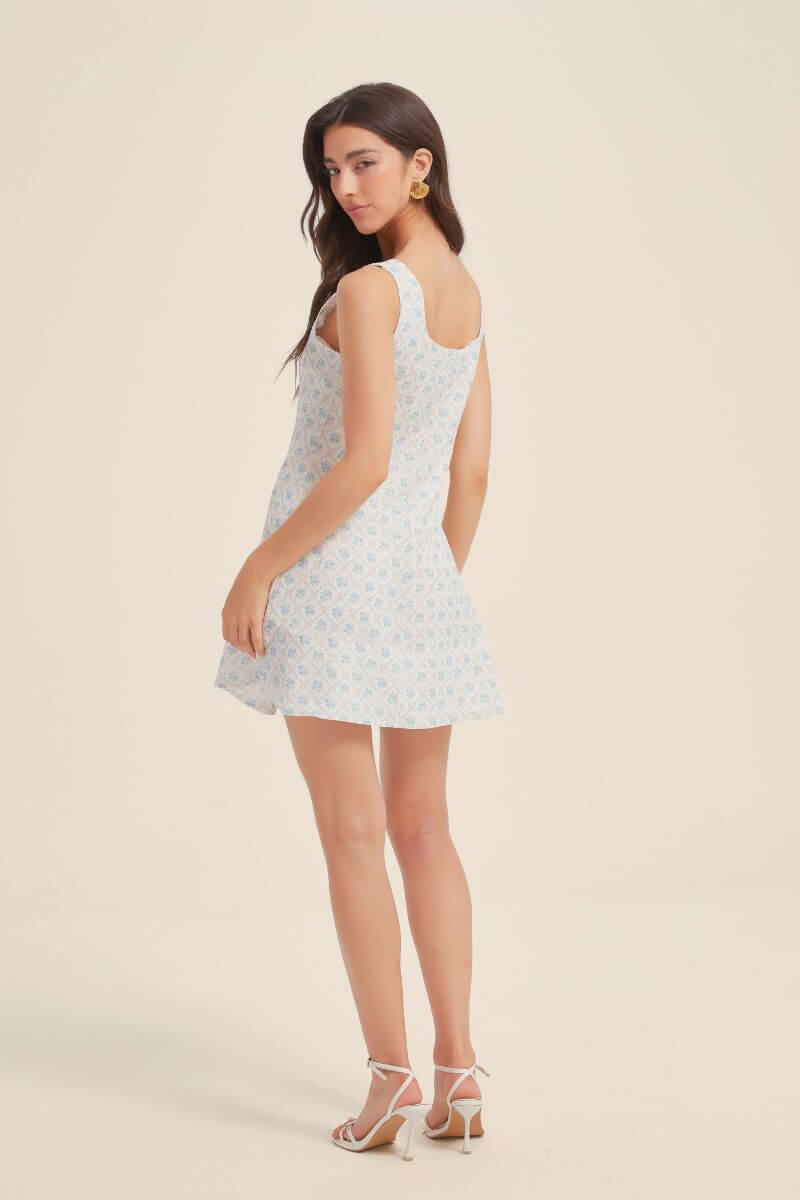 Back view of a woman wearing a blue jacquard floral mini dress with wide straps and a subtle flare skirt, styled with heels.