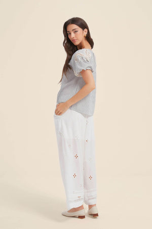 Side view of a woman wearing a blue striped blouse with puff sleeves, lace shoulder details, and relaxed fit, styled with white embroidered wide-leg pants and cream flats.