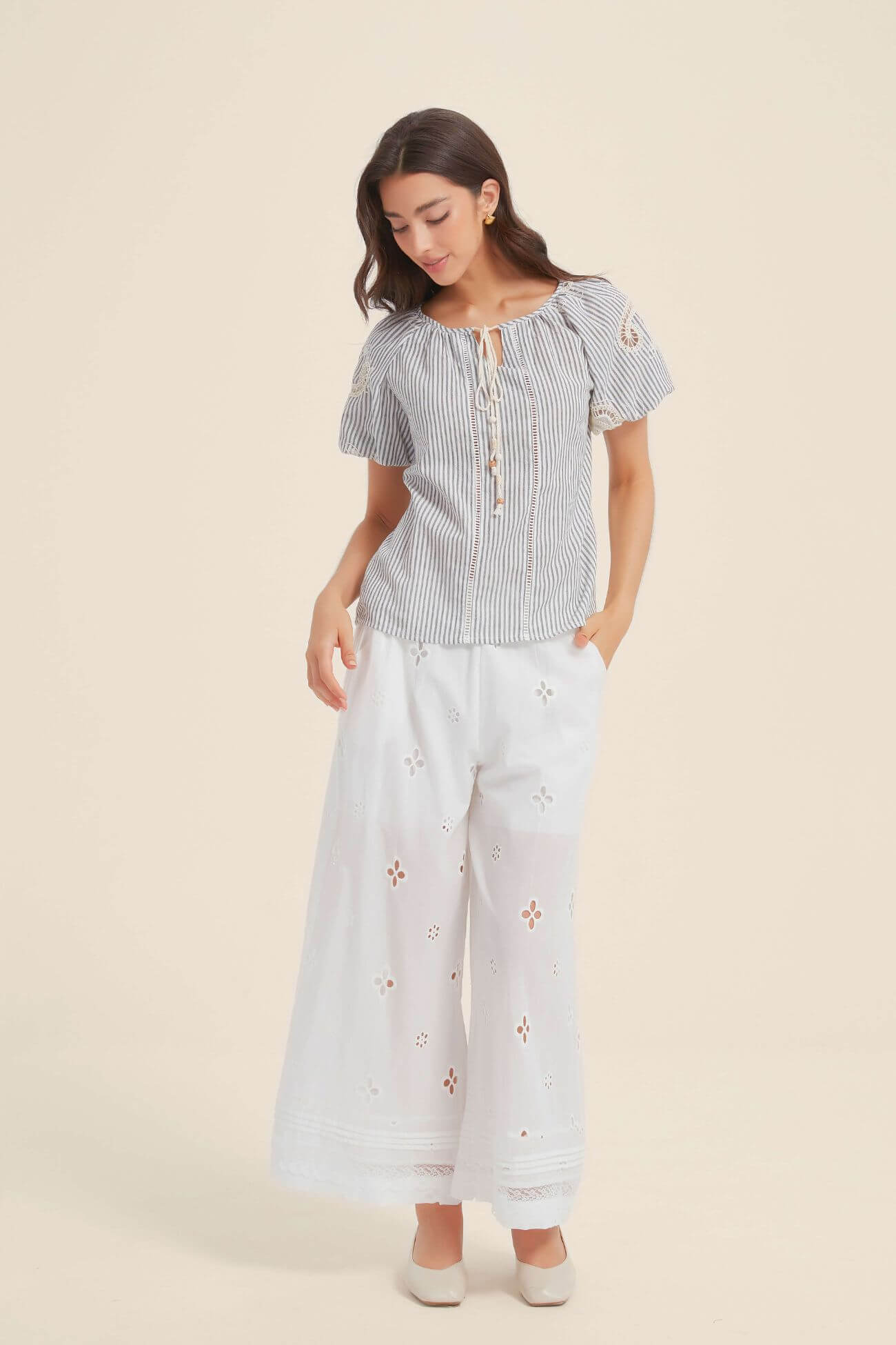 Full outfit view of a woman in a blue and white striped lace-trim blouse with tie-front tassels, paired with white eyelet wide-leg pants and cream shoes for a soft summer look.