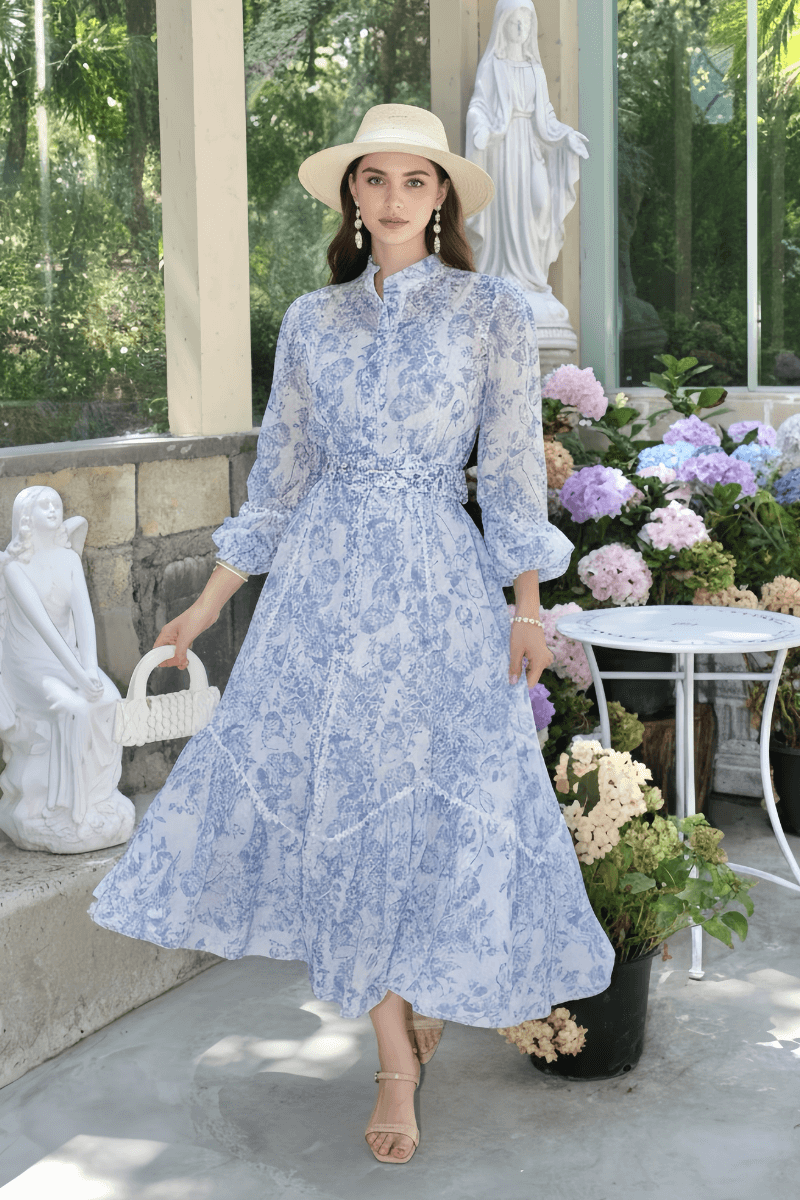 A woman in a light blue toile print puff-sleeve maxi dress with a belted waist, accessorized with a straw hat and pearl earrings, standing among hydrangeas and statues.