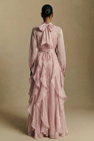 Back view of a blush pink chiffon maxi dress showing a soft bow tie at the neckline, gathered waist, and layered ruffle skirt flowing to the floor.