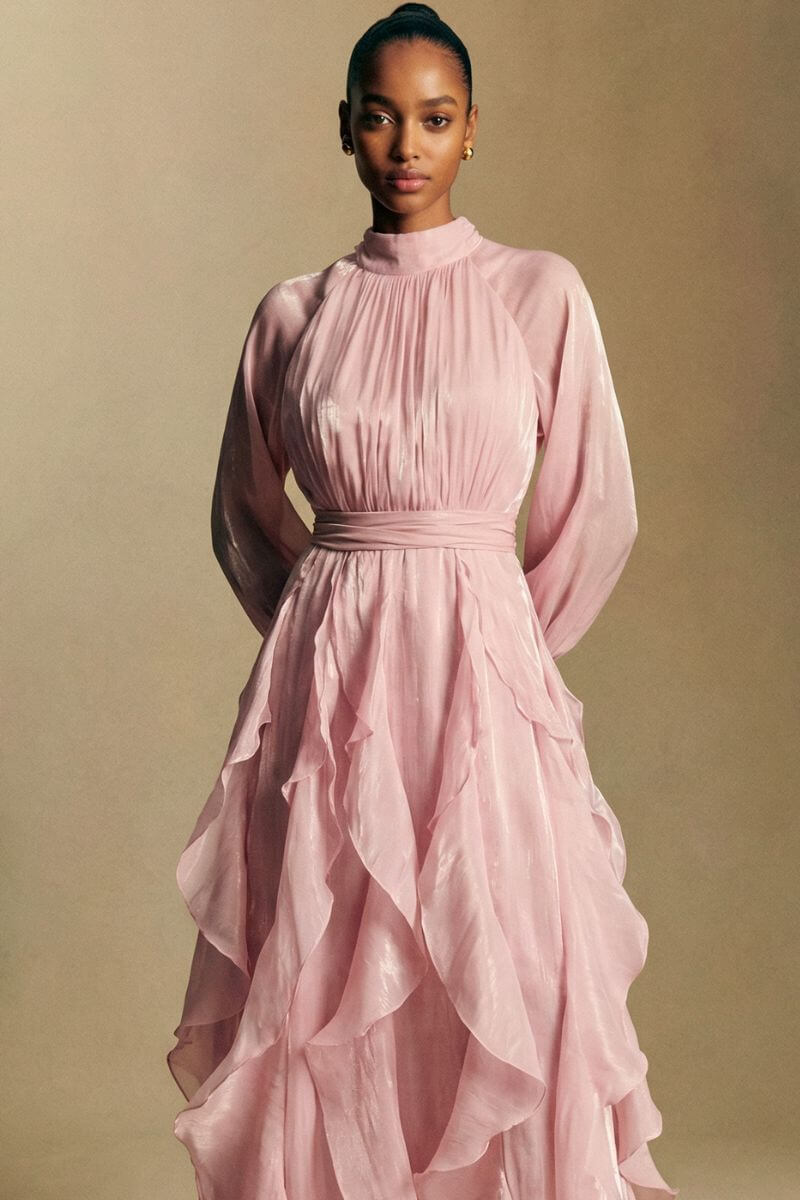 Back view of a blush pink chiffon maxi dress showing a soft bow tie at the neckline, gathered waist, and layered ruffle skirt flowing to the floor.