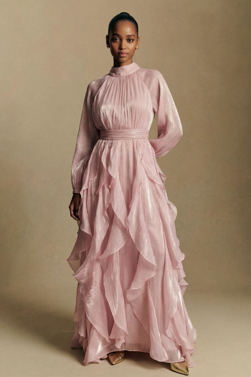 A model stands facing forward wearing a blush pink chiffon maxi dress featuring a gathered high neck, cinched waist, and cascading ruffle layers for an elegant silhouette.