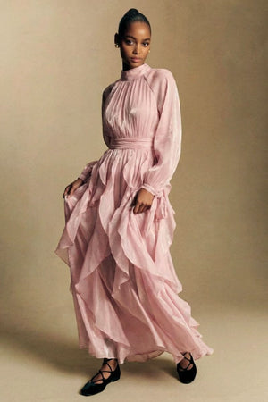 A model walks gracefully in a blush pink chiffon maxi dress with soft ruffles, high neckline, and flowing layered skirt against a warm neutral studio backdrop.