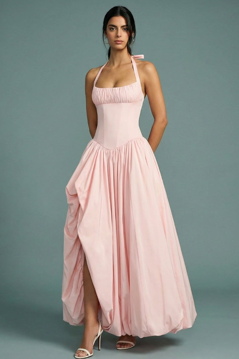A model wears a blush pink halter corset maxi dress featuring a fitted bodice, softly gathered full skirt, and fluid drape, styled with delicate heels in a minimal studio setting.