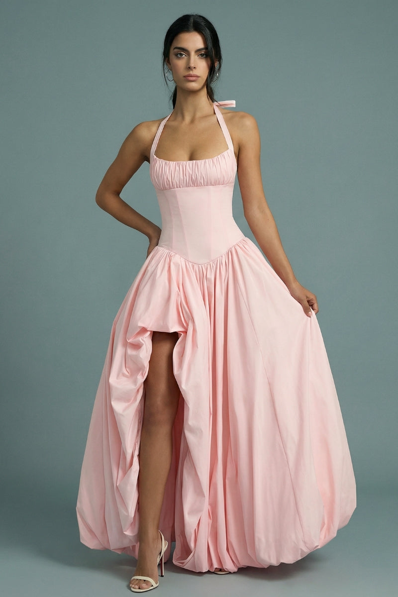 A model poses in a blush pink halter corset maxi dress with a softly structured bodice, dramatic gathered skirt, and a subtle front slit that reveals movement and volume.