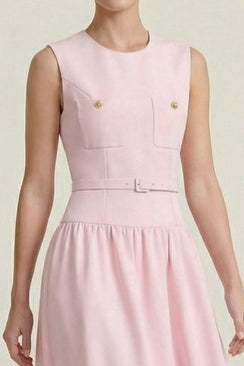 Close-up of a blush pink sleeveless dress showing the structured bodice with gold-button flap pockets and a matching slim belt in a minimal neutral studio background.