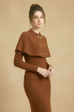 A woman poses in a brown midi cape sweater dress with structured shoulders and gold button accents, looking slightly to the side in a beige studio.