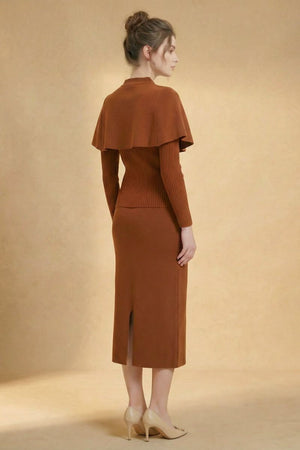 A woman stands facing away, wearing a brown knit cape-shoulder sweater dress with a ribbed bodice and back slit, styled with nude heels in a neutral studio setting.