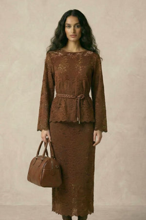A woman in a brown lace two-piece set featuring a long-sleeve lace top, a matching midi skirt, and a braided belt cinching the waist.