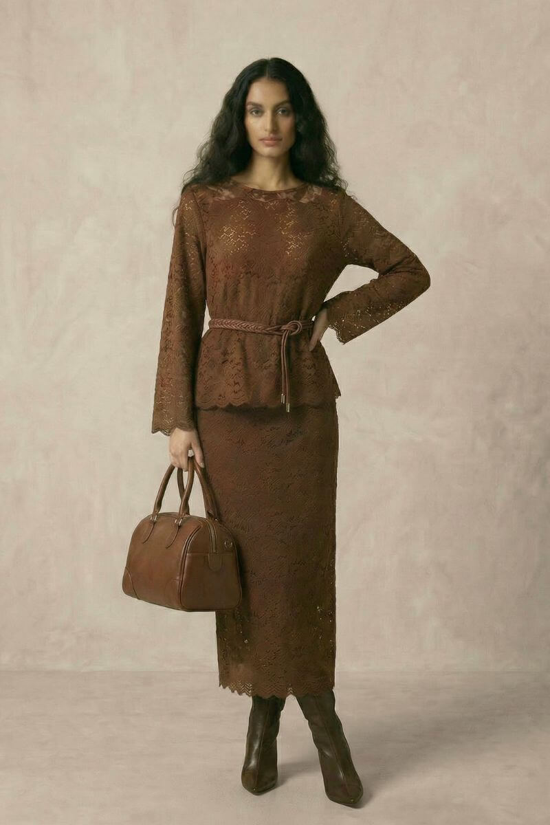 A woman wearing a brown lace top and matching midi skirt styled with a braided waist belt, brown heeled boots, and a structured leather handbag.