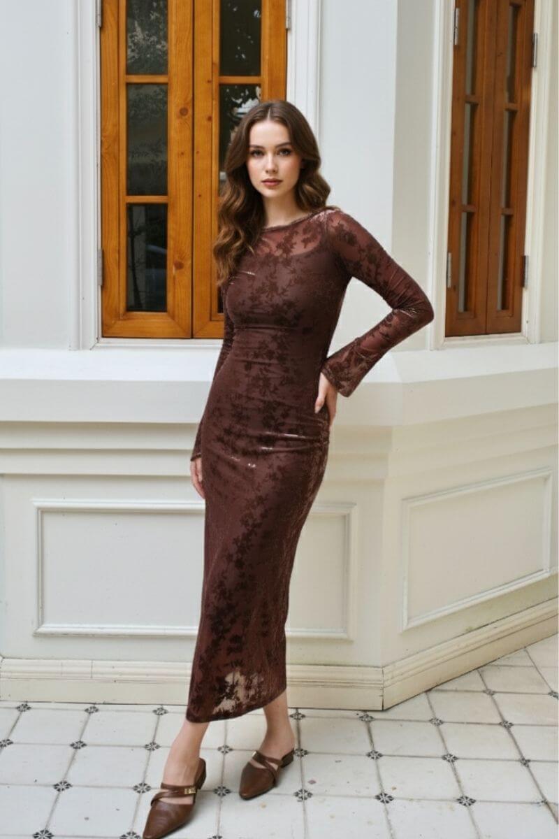 Model posing indoors against white paneled walls wearing a brown sheer velvet floral bodycon midi dress with long sleeves and boat neckline, styled with brown pointed-toe flats for an elegant autumn look.