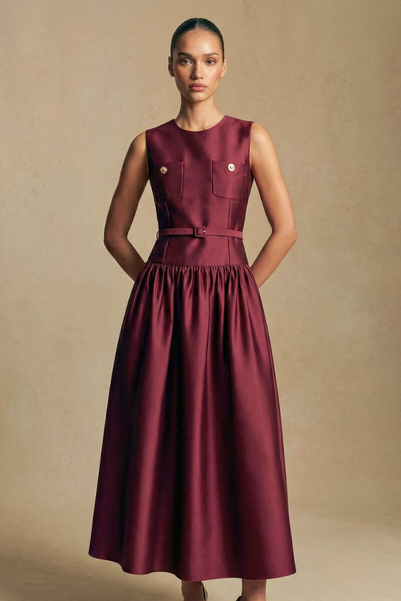 Model stands facing forward in a burgundy sleeveless midi dress with structured bodice, decorative buttons, slim belt, and elegant mid-length skirt.