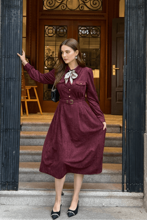 Model posing at the entrance of a building, wearing a burgundy belted midi dress with a bow scarf and black pointed heels.