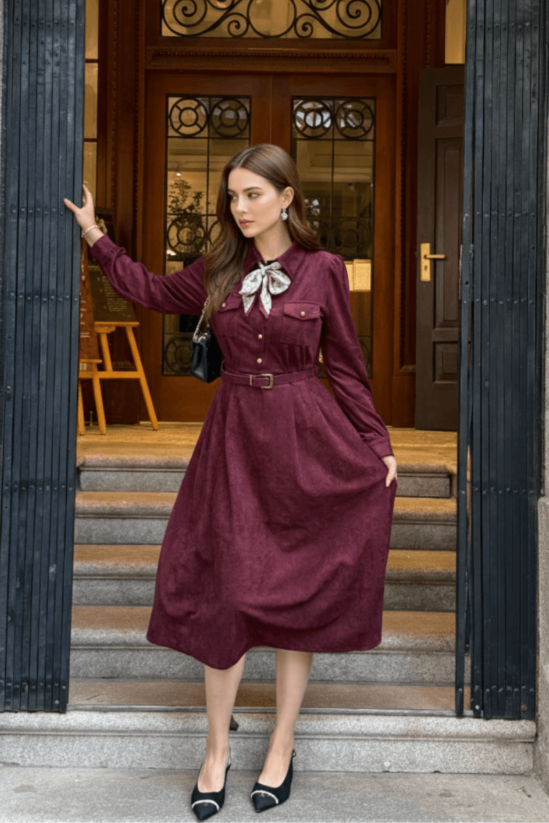 Model posing at the entrance of a building, wearing a burgundy belted midi dress with a bow scarf and black pointed heels.