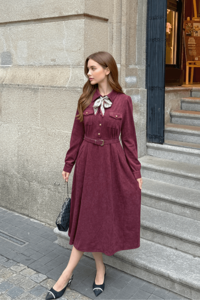 Model wearing a burgundy belted midi dress with a decorative bow scarf, styled with black heels and a quilted handbag on a city street.