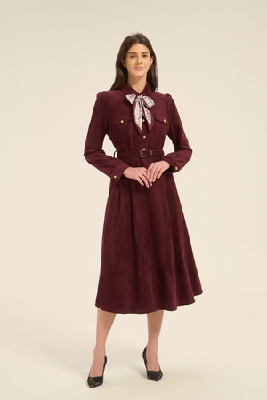 Model facing forward in a burgundy midi dress with long sleeves, gold button details, a belted waist, and a patterned bow scarf tied at the collar.