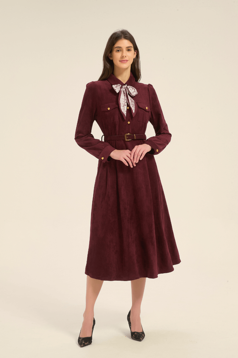 Model facing forward in a burgundy midi dress with long sleeves, gold button details, a belted waist, and a patterned bow scarf tied at the collar.