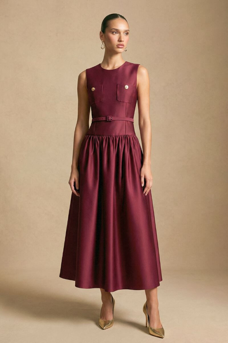Model wears a burgundy sleeveless midi dress with a tailored waist, subtle sheen fabric, and softly flared skirt, styled with pointed heels in a minimal studio scene.