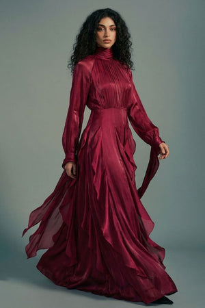 A model wears a burgundy high-neck maxi dress with long sleeves and cascading ruffle layers, styled with soft movement against a muted studio background.