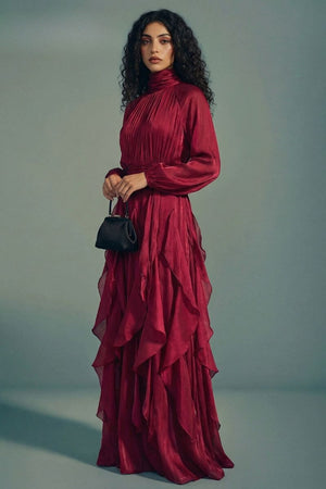 A front-facing view of a burgundy high-neck long sleeve maxi dress featuring a softly pleated bodice and layered ruffle skirt, styled with a structured mini handbag.